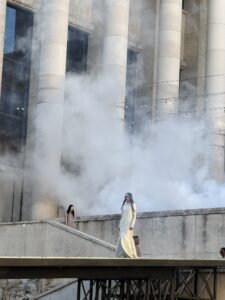 A model in a long pale gown walks a raised runway in front of the colonnade of the Palais de Tokyo as fog drifts across the stone columns.