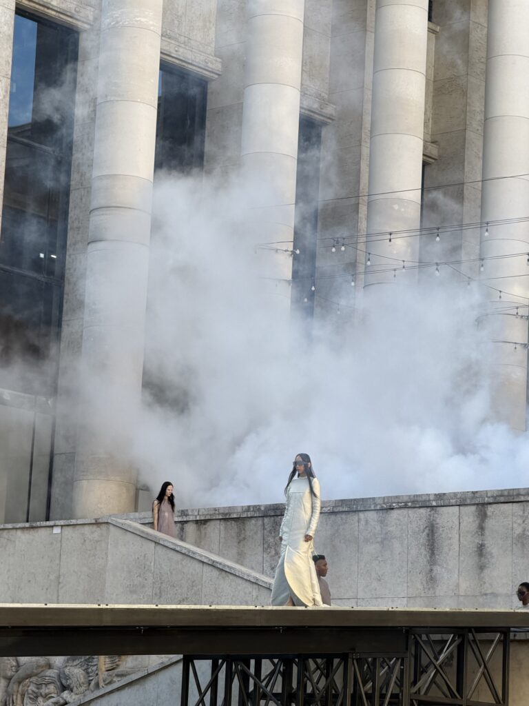 A model in a long pale gown walks a raised runway in front of the colonnade of the Palais de Tokyo as fog drifts across the stone columns.