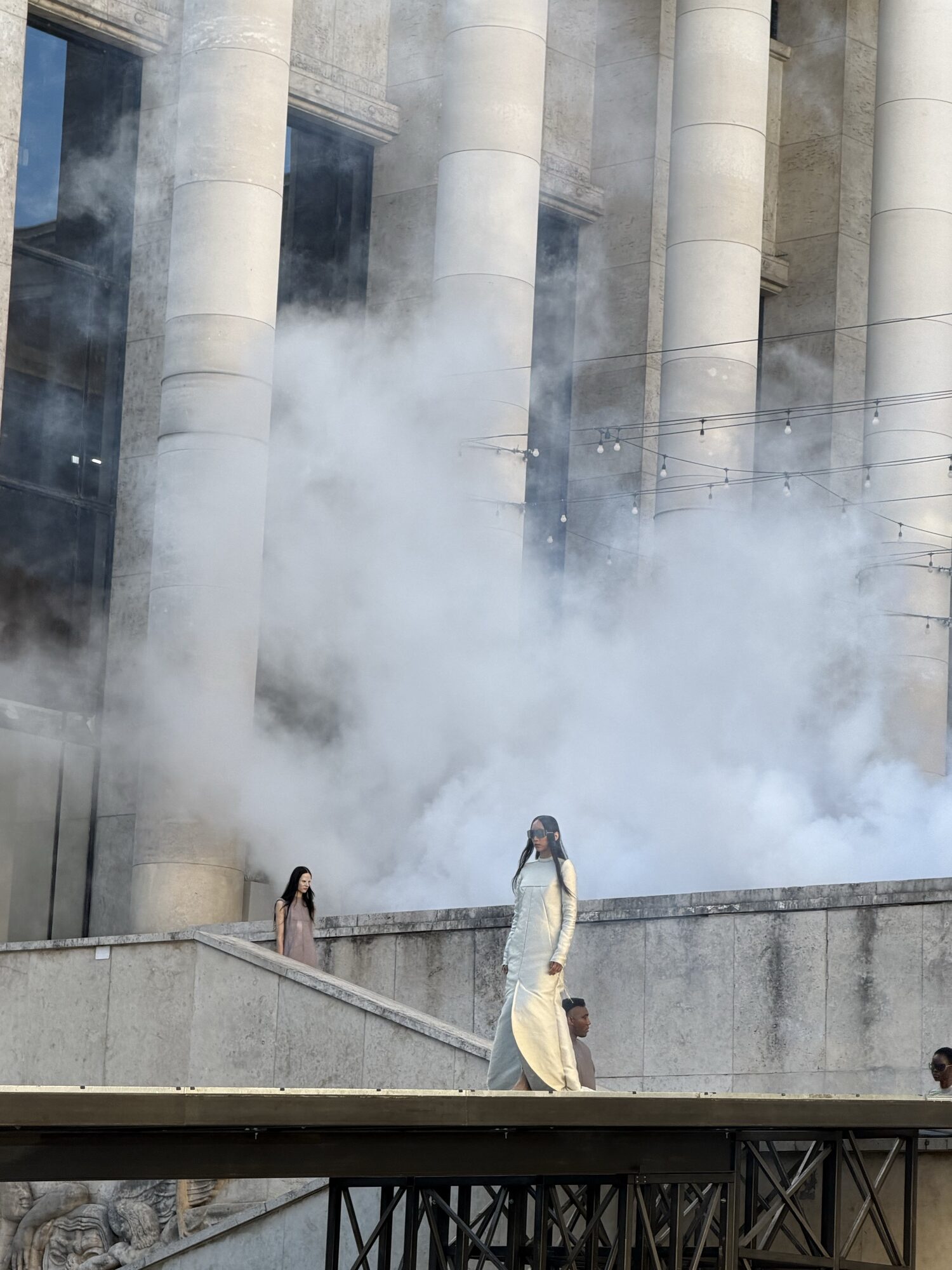 A model in a long pale gown walks a raised runway in front of the colonnade of the Palais de Tokyo as fog drifts across the stone columns.
