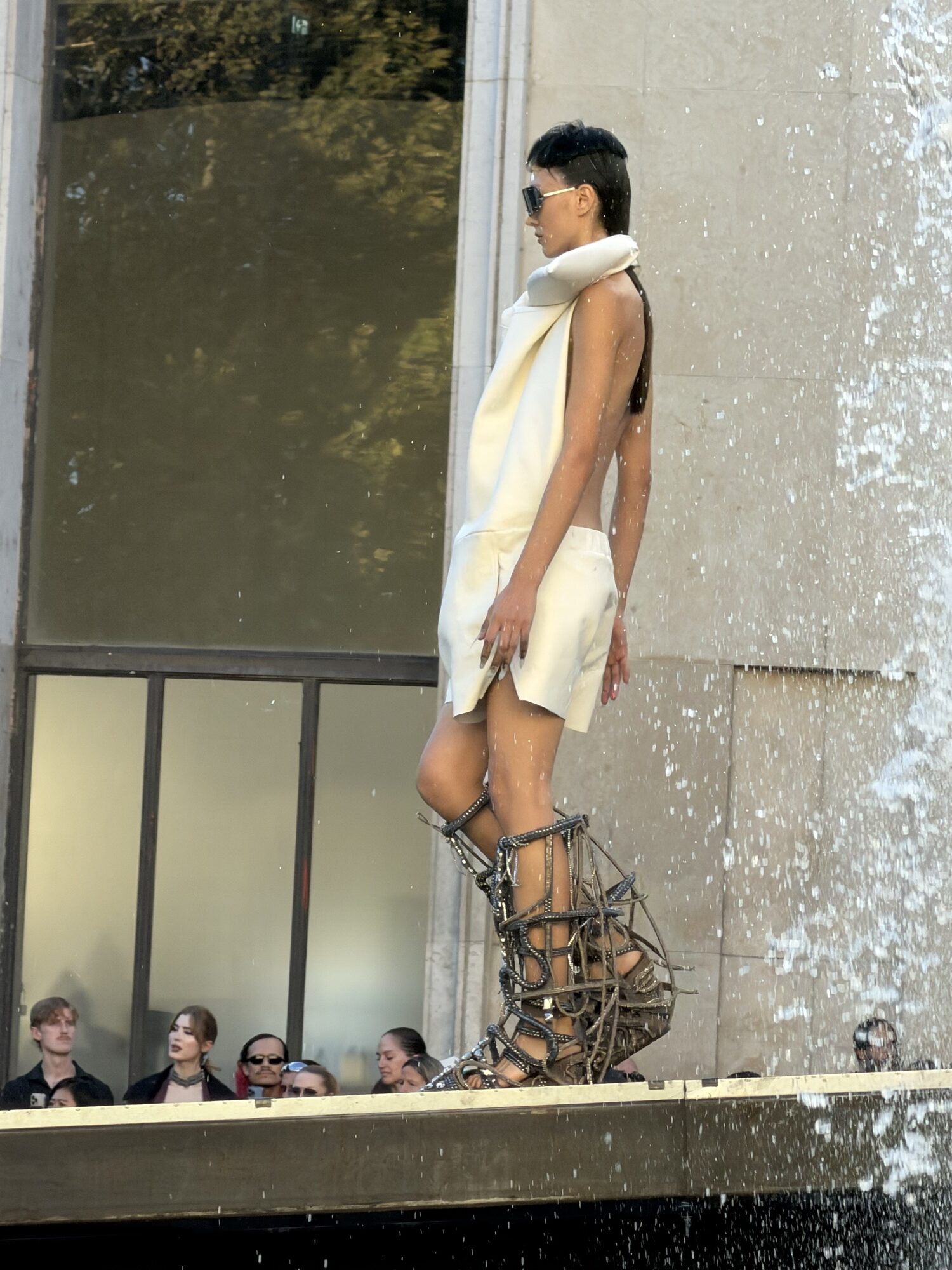 A model in a cream sleeveless tunic and shorts with tall caged metal boots walks beside a fountain, water spraying across the runway.