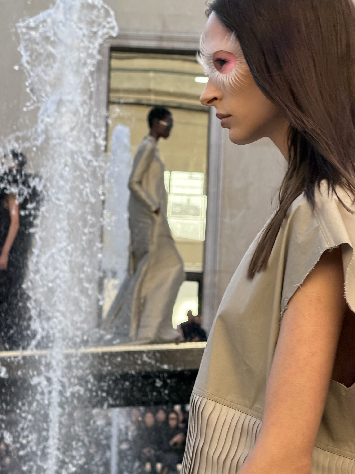 A model in a raw-edged khaki vest and pleated skirt stands in profile with pink eye makeup, a fountain and another model behind her.
