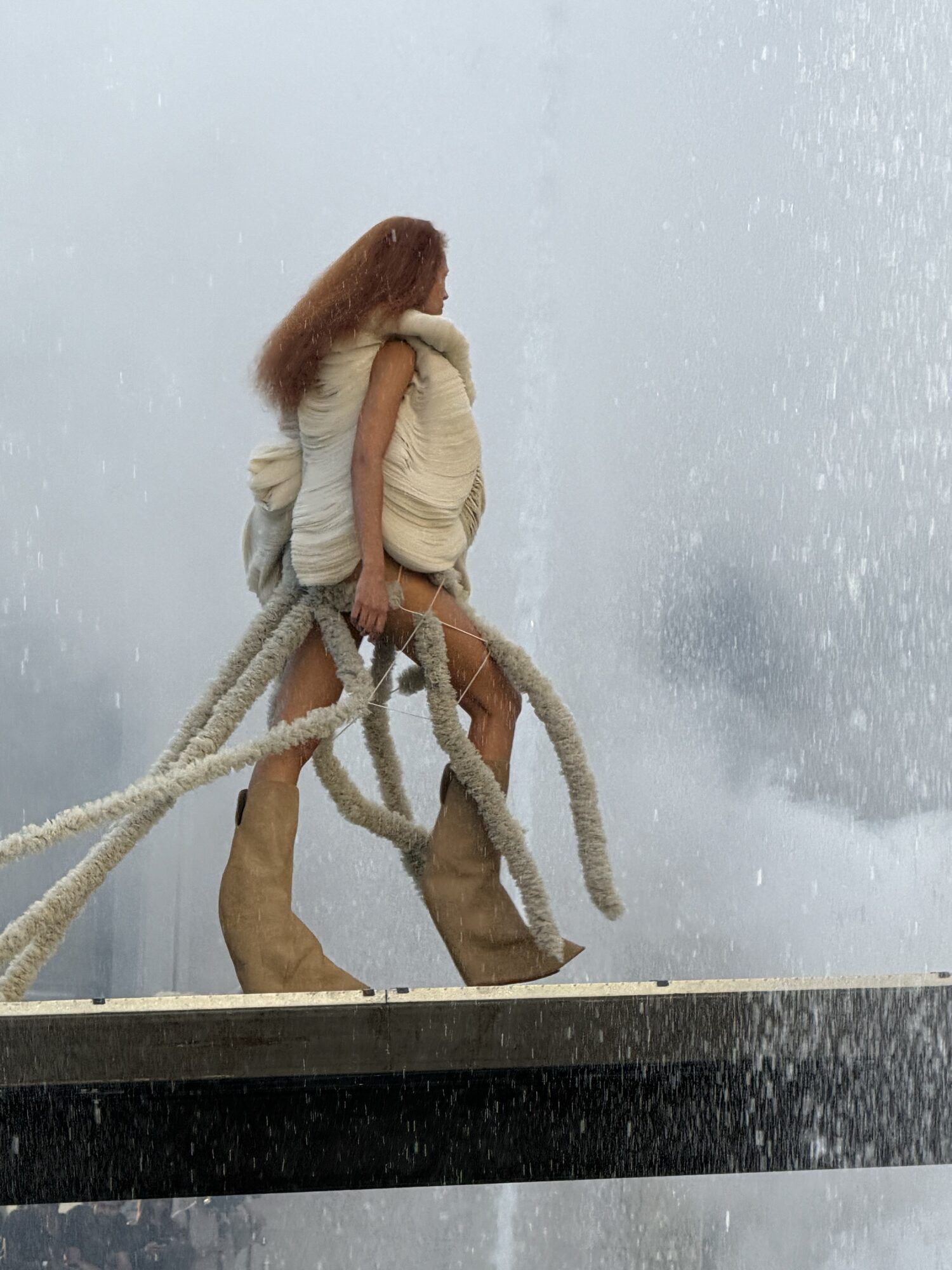 A model in a draped cream dress trailing long ropes of fringe walks through heavy fountain spray on the runway.
