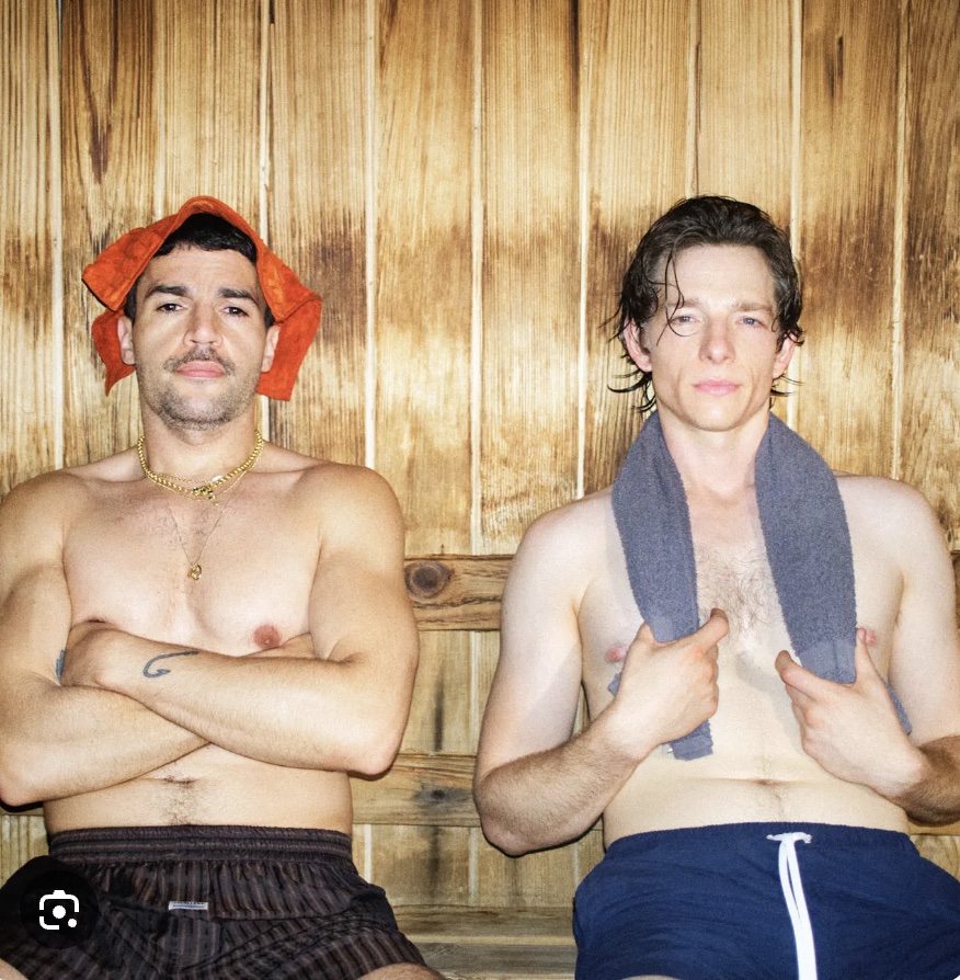 Two shirtless men sit side by side on a wooden sauna bench, one with an orange towel draped over his head.