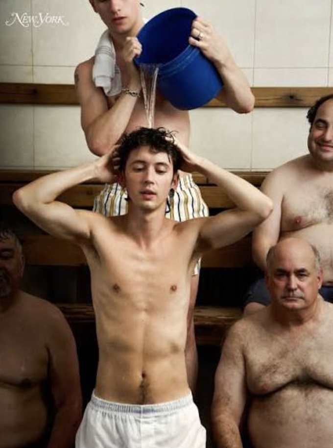 A young man stands shirtless in a bathhouse while another pours water from a blue bucket over his head, older bathers seated on either side.