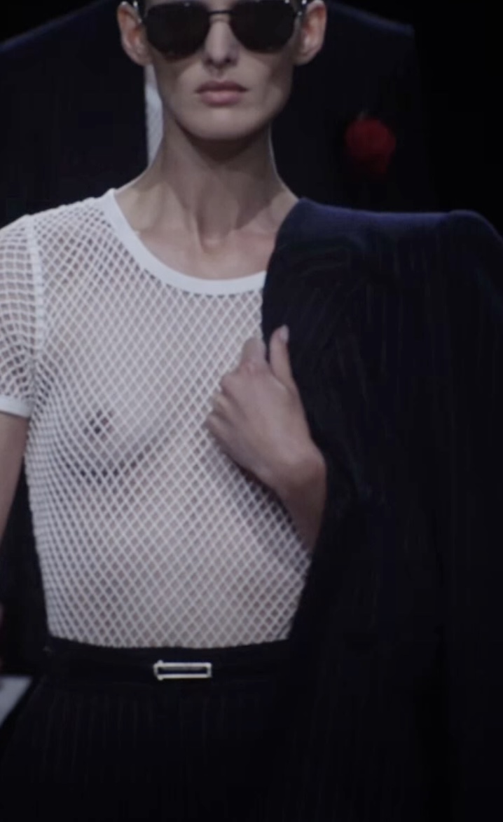 Close-up of a model on a dark runway in a white open-weave mesh top, holding a navy pinstriped jacket across one shoulder.