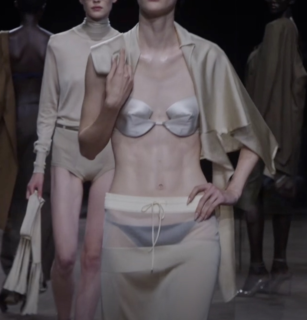 Models on a dim runway in pale beige and cream; the foreground look pairs a silver bra with a sheer low-slung skirt and a draped jacket.